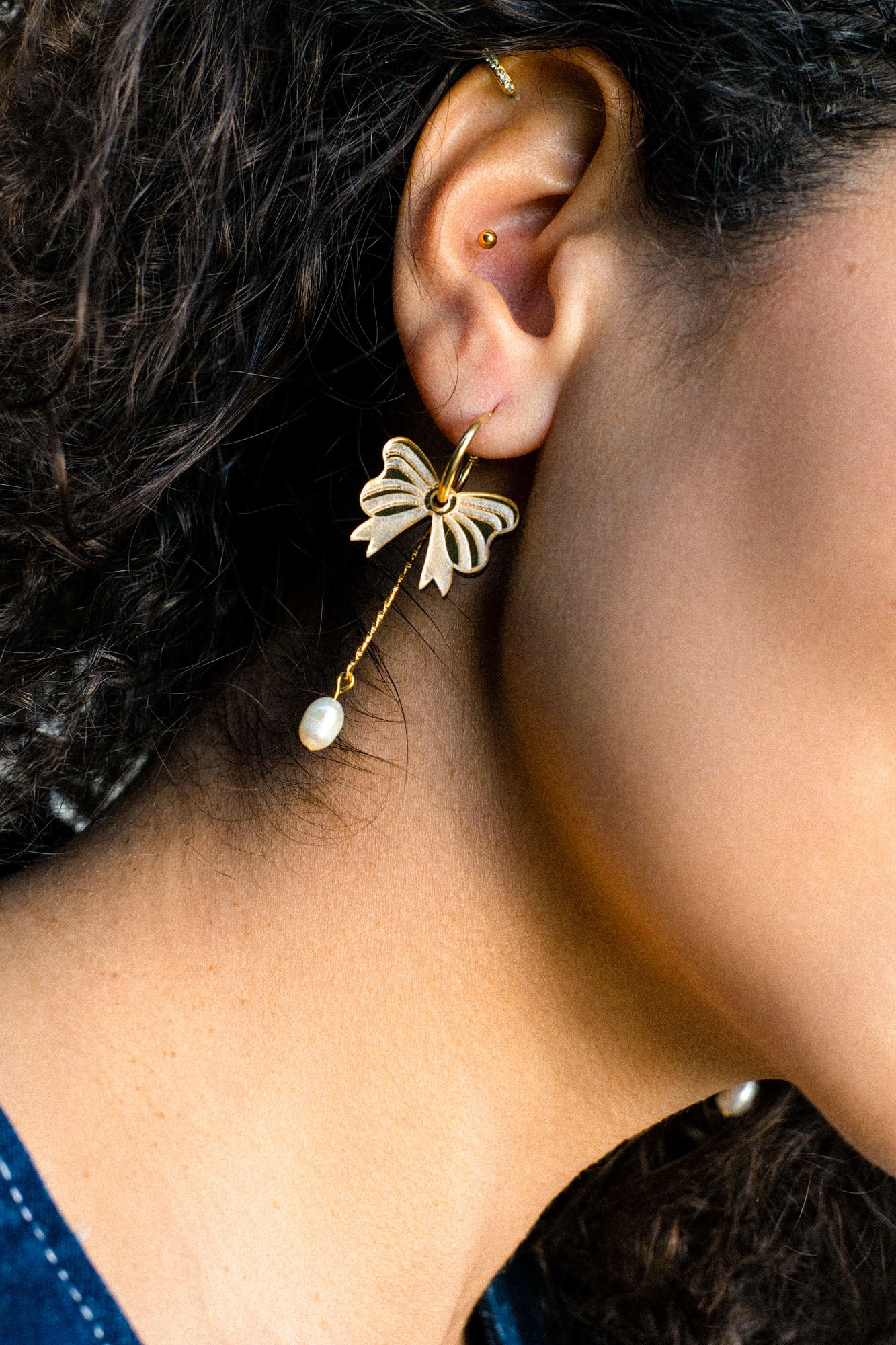 Close-up of a person's ear adorned with a gold-plated butterfly earring featuring a dangling pearl, reminiscent of elegant freshwater pearl earrings. The person has curly dark hair and a second small stud in the upper ear cartilage.