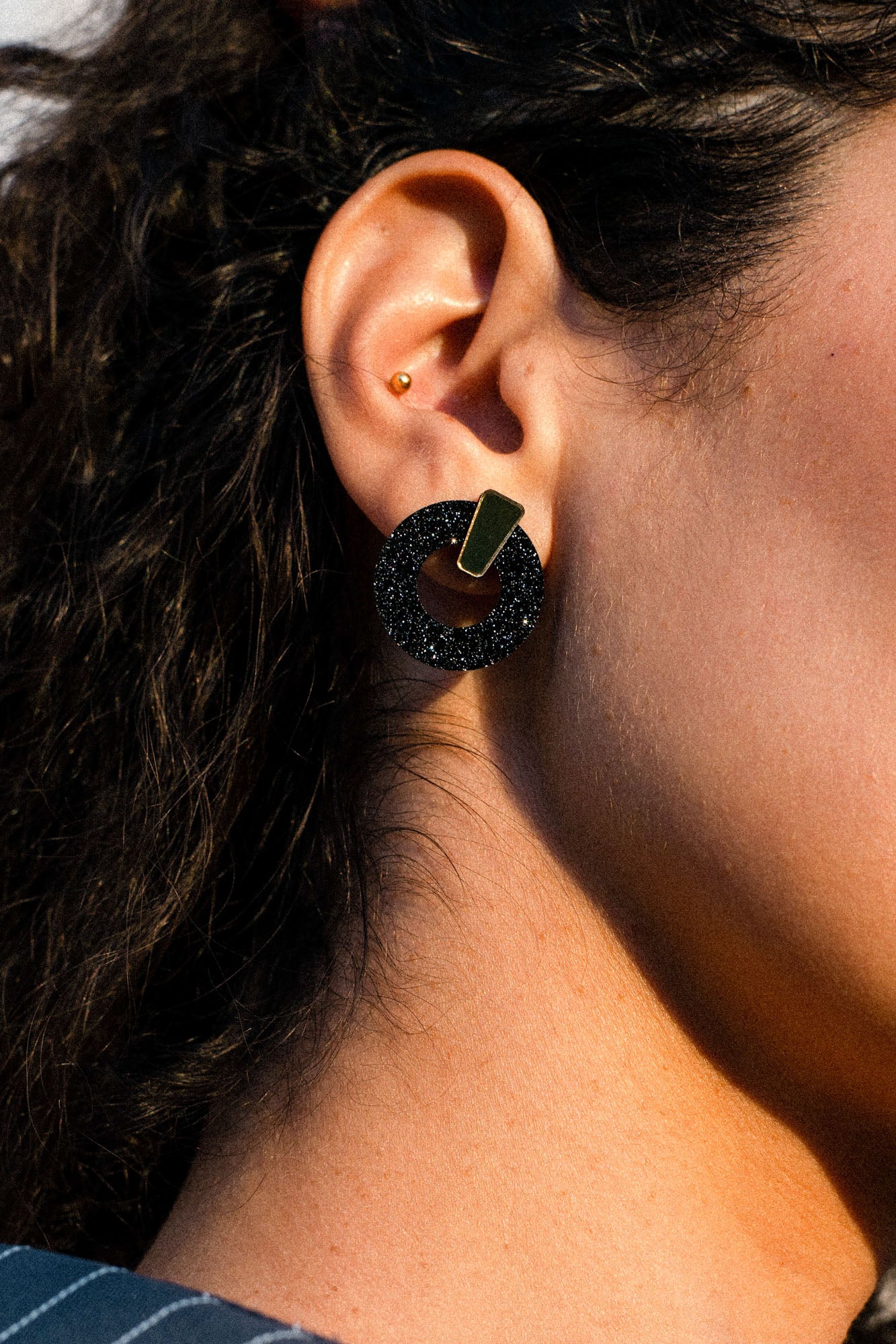 Close-up of a person’s ear with two earrings: a small gold stud in the upper ear and a large black circular earring with a gold accent in the earlobe. The black earring, crafted in an acrylic glass finish, adds a modern touch to the look. The person has curly dark hair.