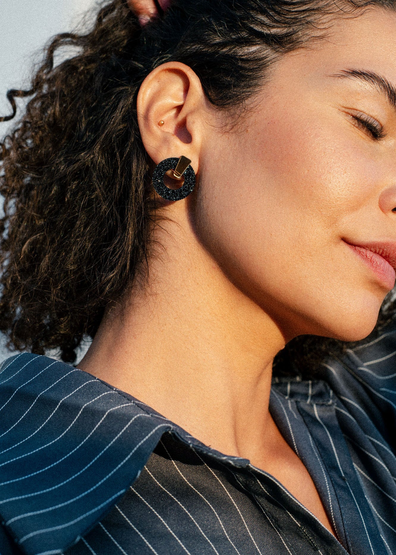 A woman with curly hair wears a dark, geometric acrylic glass earring and a pinstriped shirt. The image shows her profile and focuses on her ear and jawline, with sunlight highlighting her skin.