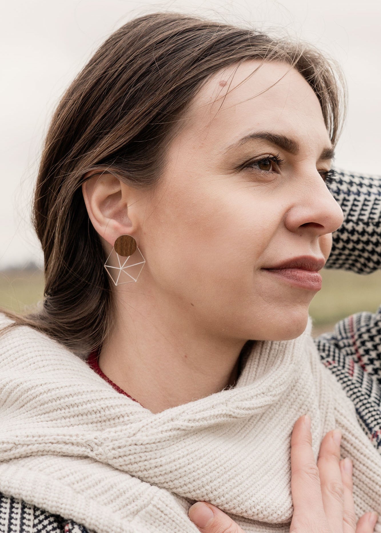 A woman in a houndstooth coat and cream scarf, embodying minimalist elegance, stands outdoors on a grassy, overcast day. She gazes thoughtfully into the distance, one hand on her chest and the other behind her head.