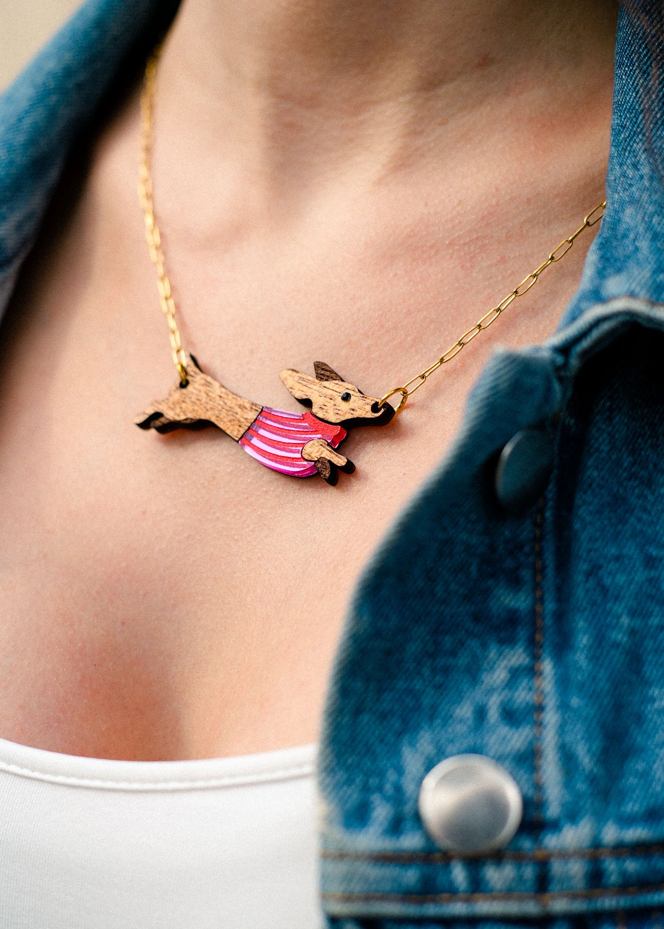 A close-up of a person wearing a gold necklace with a handcrafted Dachshund pendant in a pink striped sweater, made from sustainable wood. The person is also wearing a white top and a blue denim jacket.