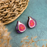 A pair of fig-shaped, hand-painted earrings with purple and pink detailing are displayed on a green surface next to a woven basket. Blurred beige plant stems are visible in the foreground.
