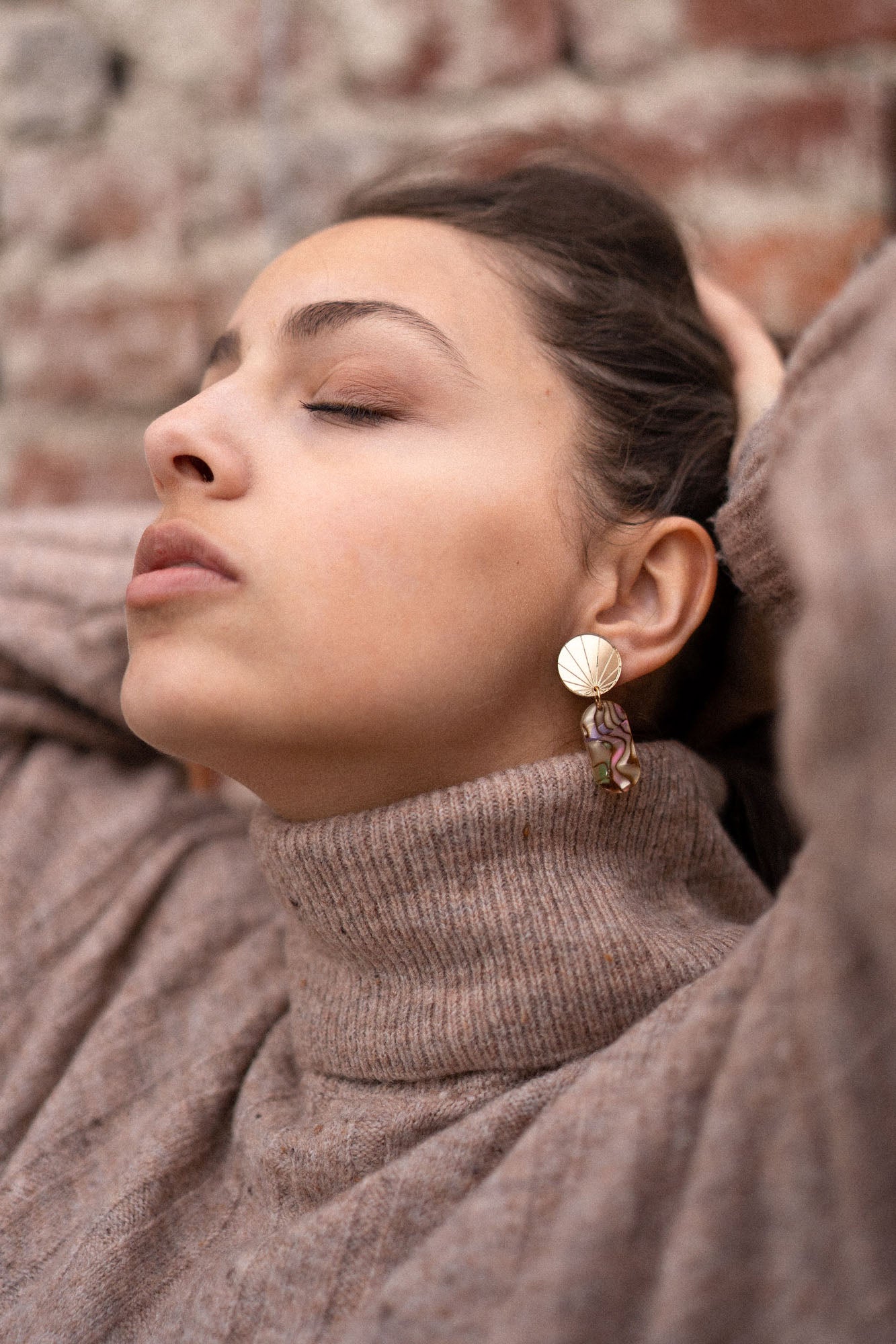 A woman wearing a beige turtleneck sweater and handcrafted gold earrings poses with her eyes closed, head tilted back, and hands resting behind her head in front of a brick wall.
