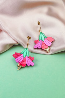 Handcrafted Hibiscus Earrings in a tropical statement style, featuring colorful pink and red petals, green leaves, and gold accents, are displayed on soft pink fabric and a mint green background.
