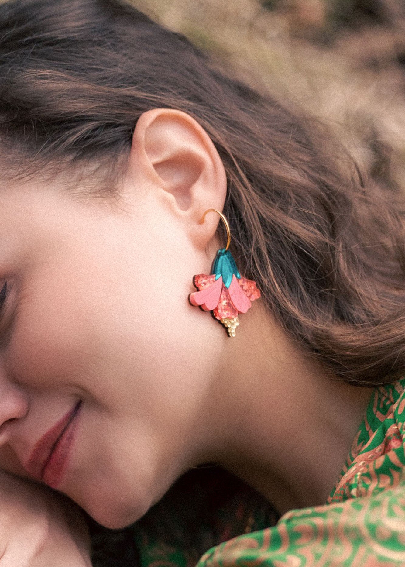 A woman with wavy brown hair wears vibrant Hibiscus Earrings—gold hoops adorned with pink, teal, and gold floral charms. She is smiling softly, her tropical statement green patterned clothing visible.