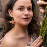 A woman with wavy brown hair and floral handcrafted Hibiscus Earrings stands in a forest, wearing an off-shoulder beige top. She gently holds a mossy tree trunk and looks at the camera with a soft, calm expression.