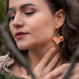 A woman with wavy brown hair and handcrafted Hibiscus Earrings gently touches her neck while standing in a forest. Her eyes are closed, branches cross before her, and she wears a beige top with ruffled details.
