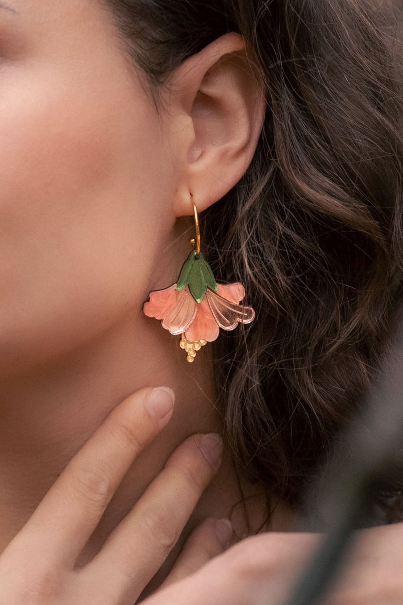 Close-up of a woman with wavy brown hair wearing handcrafted Hibiscus Earrings shaped like a pink and green flower, touching her neck gently with her fingers for a tropical statement look.