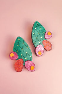 A pair of statement earrings shaped like green opuncia cactus pads with pink and orange flowers, decorated with small yellow dots, displayed on a light pink background.
