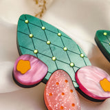 A close-up of bold statement earrings, stylized as green leaves with yellow dots and pink, orange, and purple accents—reminiscent of the opuncia cactus—displayed on soft white fabric with delicate dried flowers above.