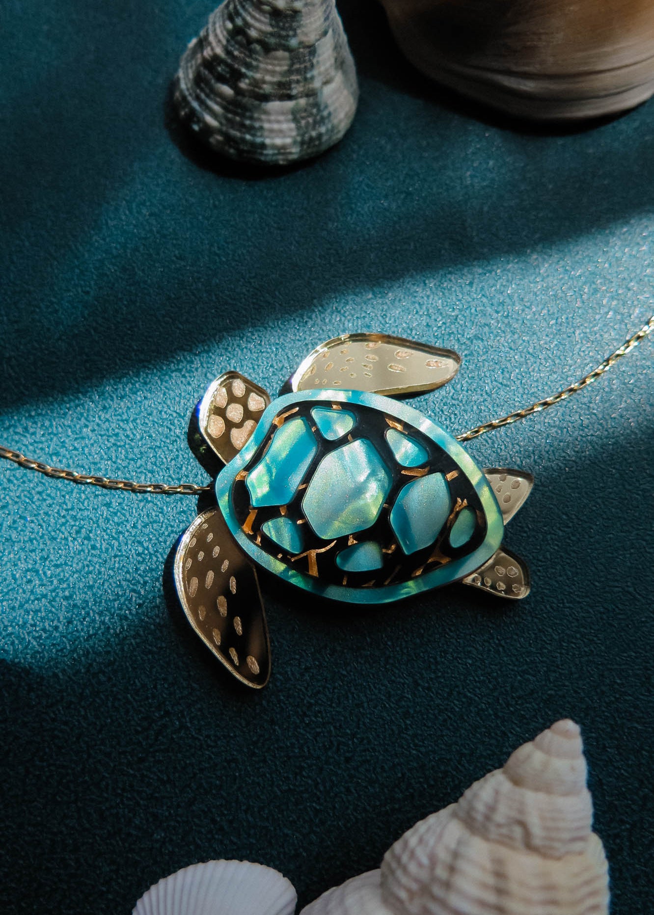 A gold and turquoise Sea Turtle Necklace pendant on a chain sits on a teal surface, surrounded by seashells and lit by soft sunlight—a stunning piece of handcrafted jewelry perfect for ocean lovers.