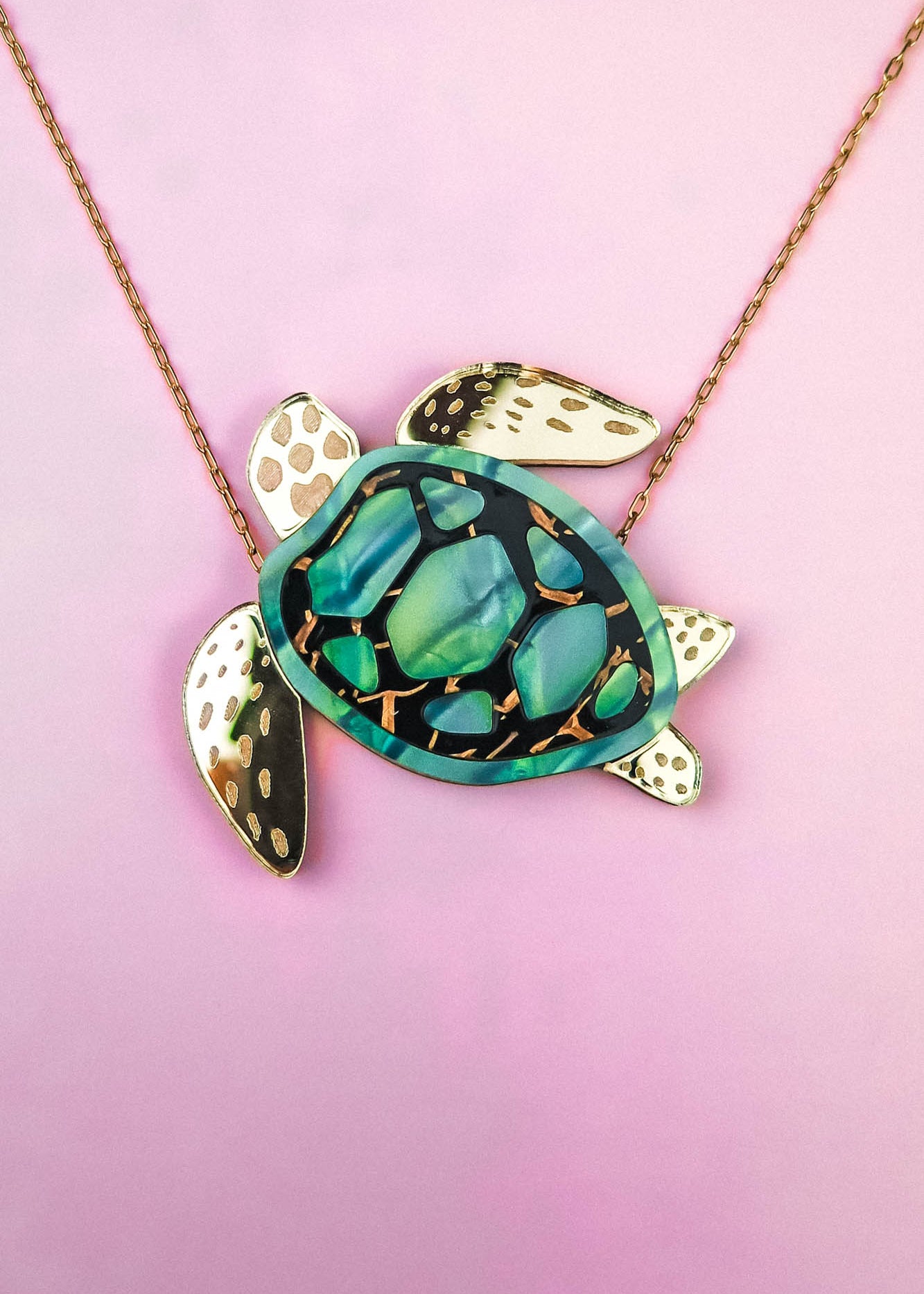 This handcrafted jewelry piece is a gold chain Sea Turtle Necklace, featuring a turtle pendant with a green and black patterned shell and elegant gold accents, beautifully set against a pink background.
