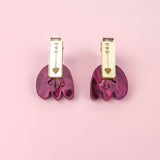 A pair of gold and purple Romantic Tulip Earrings on a pink background. The gold top section, featuring small heart engravings, perfectly complements the wavy, marbled purple acrylic bottom. These allergy-free earrings blend elegance and charm for any occasion.