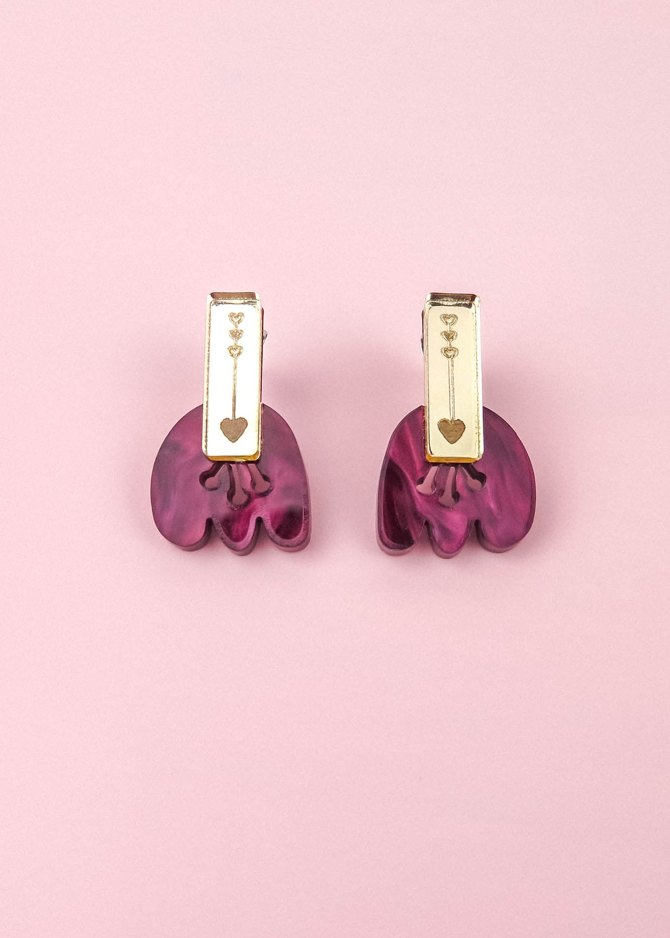 A pair of gold and purple Romantic Tulip Earrings on a pink background. The gold top section, featuring small heart engravings, perfectly complements the wavy, marbled purple acrylic bottom. These allergy-free earrings blend elegance and charm for any occasion.