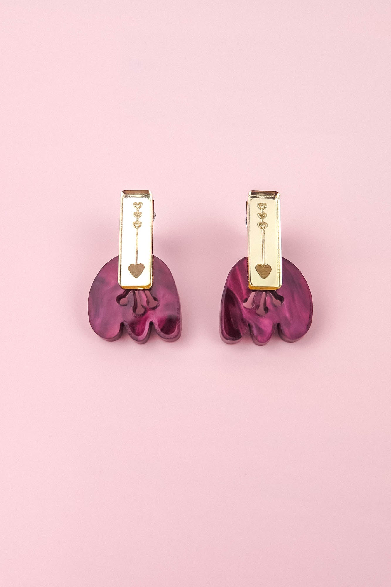 A pair of gold and purple Romantic Tulip Earrings on a pink background. The gold top section, featuring small heart engravings, perfectly complements the wavy, marbled purple acrylic bottom. These allergy-free earrings blend elegance and charm for any occasion.