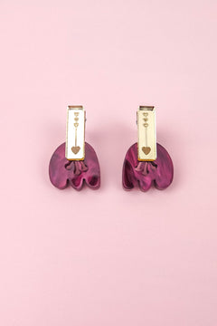 A pair of gold and purple Romantic Tulip Earrings on a pink background. The gold top section, featuring small heart engravings, perfectly complements the wavy, marbled purple acrylic bottom. These allergy-free earrings blend elegance and charm for any occasion.