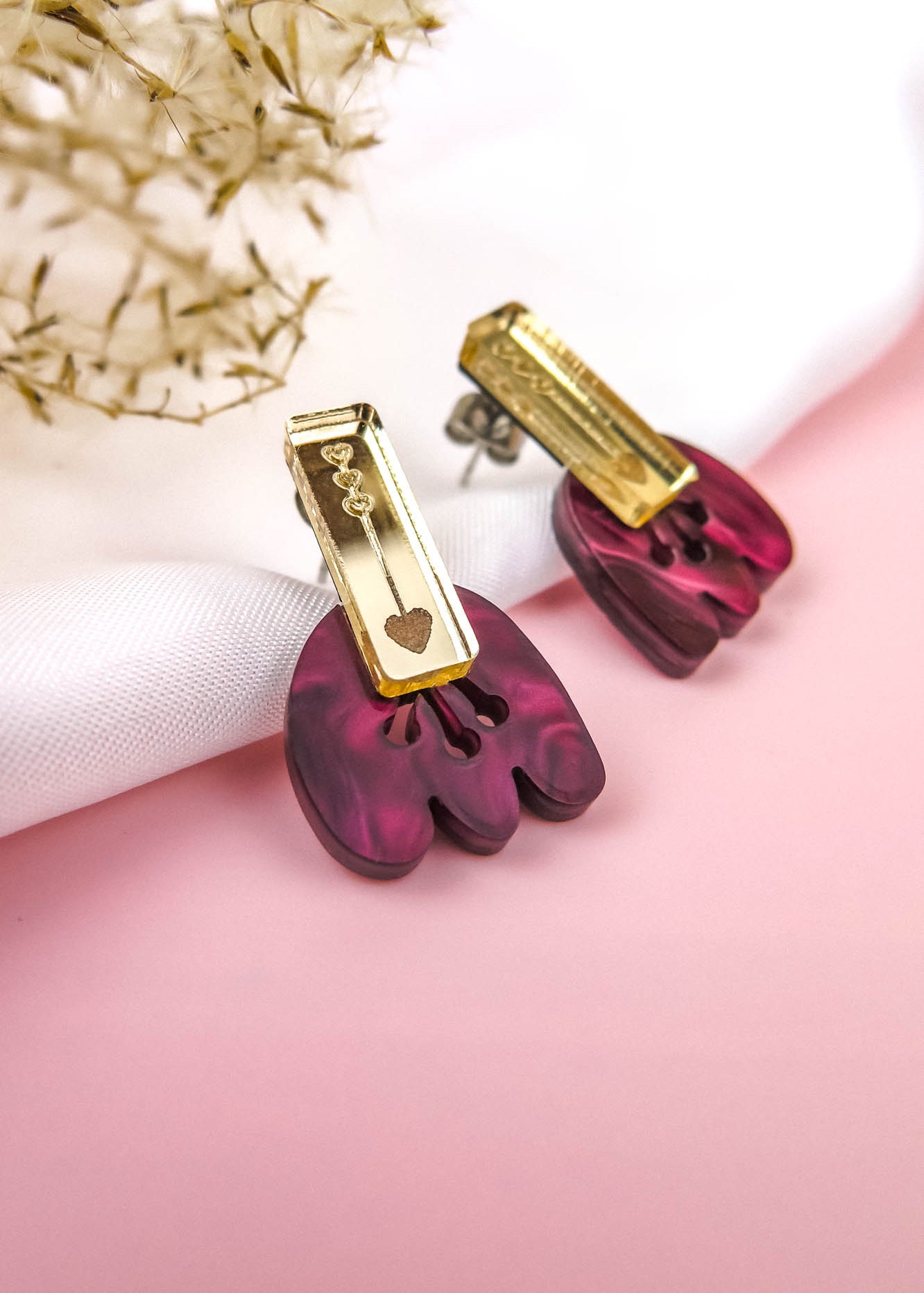 Introducing the Romantic Tulip Earrings—a pair of earrings with gold rectangular tops and dangling marbled purple acrylic shapes, featuring a small gold chain and heart charm. Allergy-free and stylish, they rest on white fabric with dried flowers on a pink background.