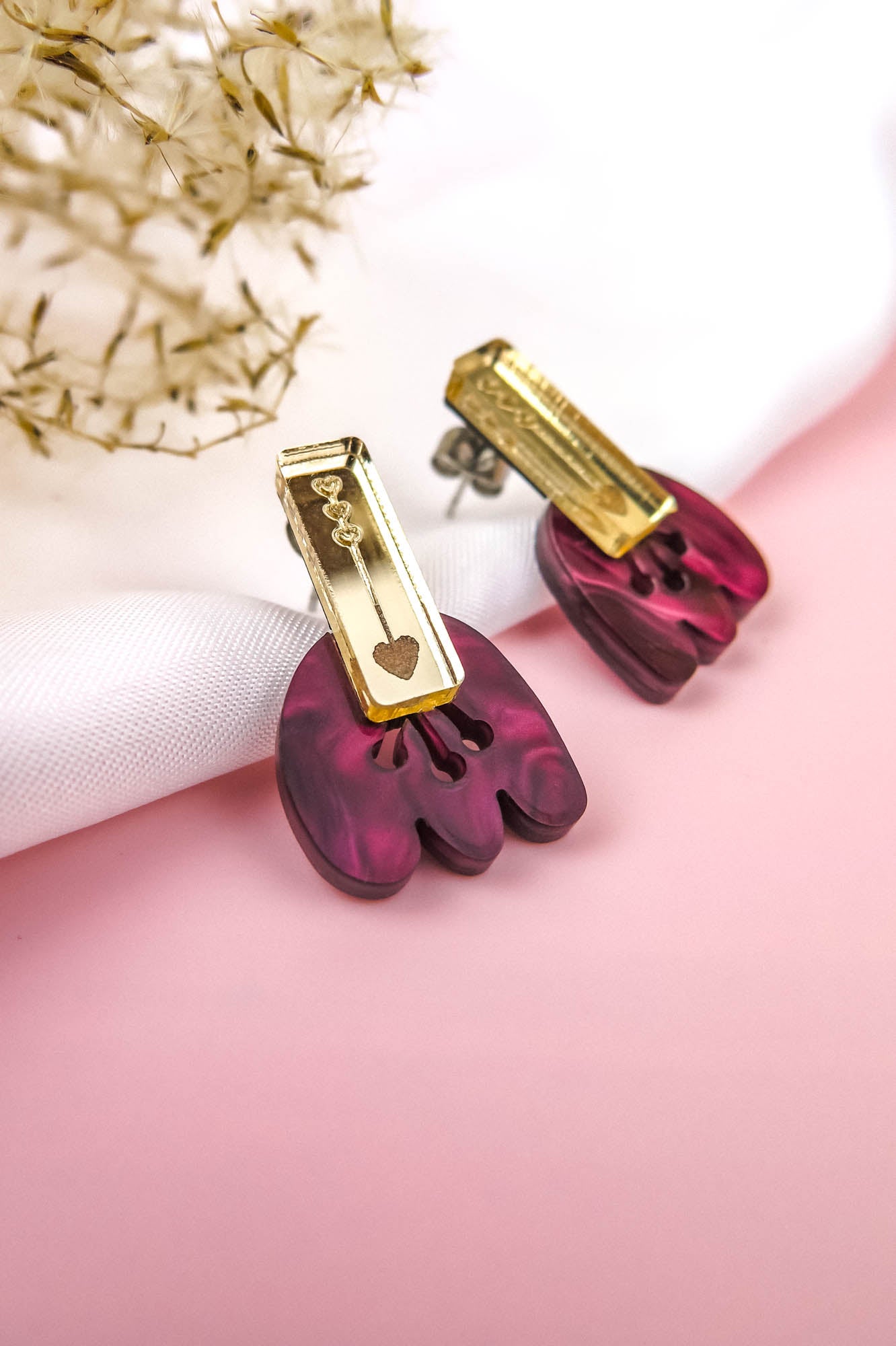 Introducing the Romantic Tulip Earrings—a pair of earrings with gold rectangular tops and dangling marbled purple acrylic shapes, featuring a small gold chain and heart charm. Allergy-free and stylish, they rest on white fabric with dried flowers on a pink background.