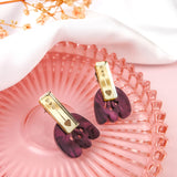 Gold and purple Romantic Tulip Earrings with heart details rest on a pink glass plate, featuring marbled purple acrylic and accented by delicate dried flowers and a white cloth in the background. These allergy-free earrings are perfect for adding a touch of whimsy to any look.