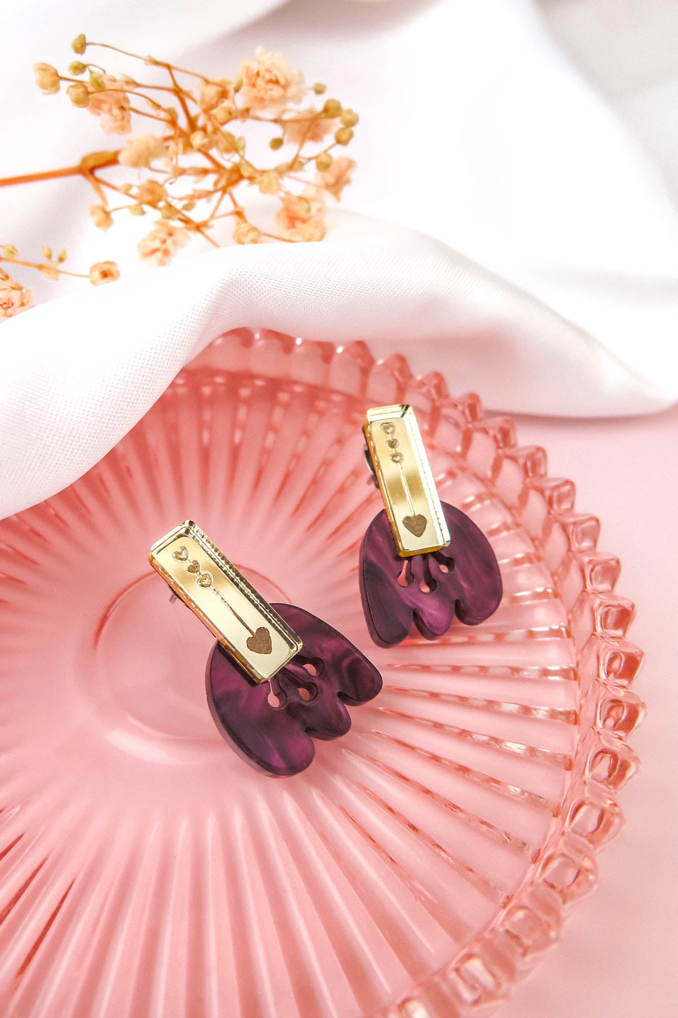 Gold and purple Romantic Tulip Earrings with heart details rest on a pink glass plate, featuring marbled purple acrylic and accented by delicate dried flowers and a white cloth in the background. These allergy-free earrings are perfect for adding a touch of whimsy to any look.