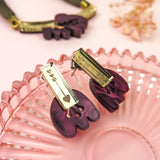 Purple and gold marbled purple acrylic earrings shaped like claws rest on a pink glass dish. A matching necklace and dried flowers are placed on a soft pink background in the distance.