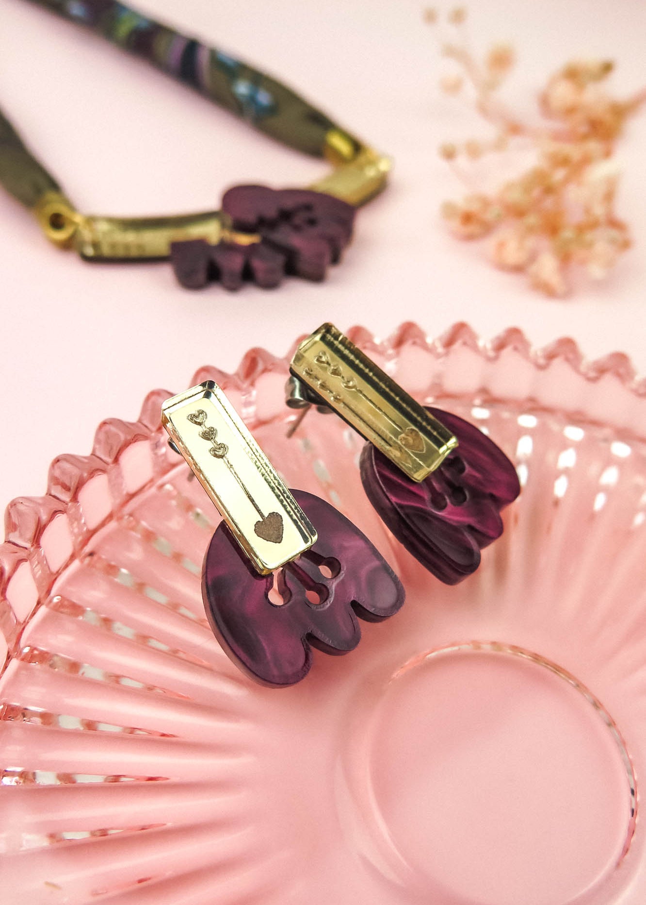 Purple and gold marbled purple acrylic earrings shaped like claws rest on a pink glass dish. A matching necklace and dried flowers are placed on a soft pink background in the distance.