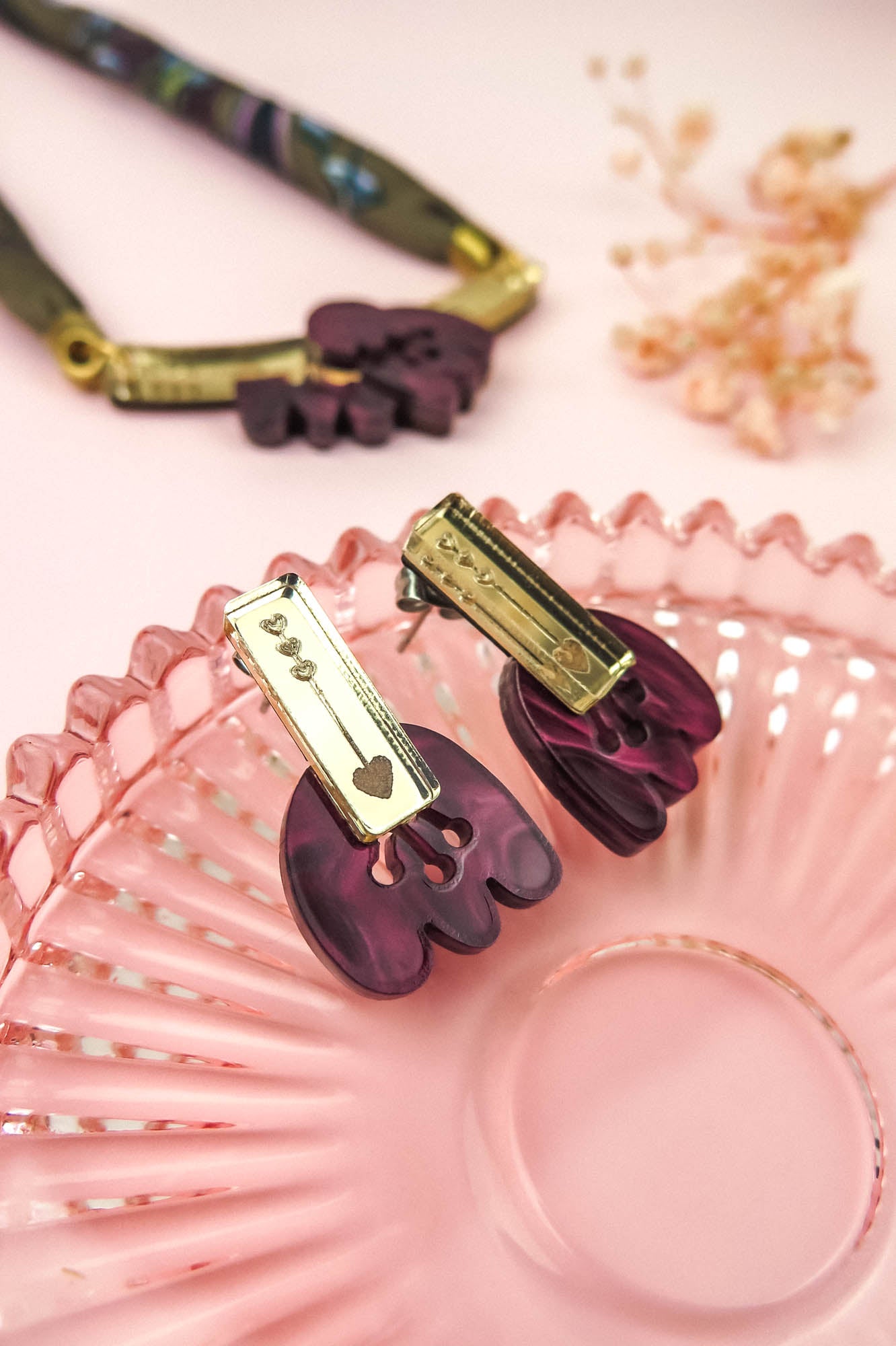 Purple and gold marbled purple acrylic earrings shaped like claws rest on a pink glass dish. A matching necklace and dried flowers are placed on a soft pink background in the distance.