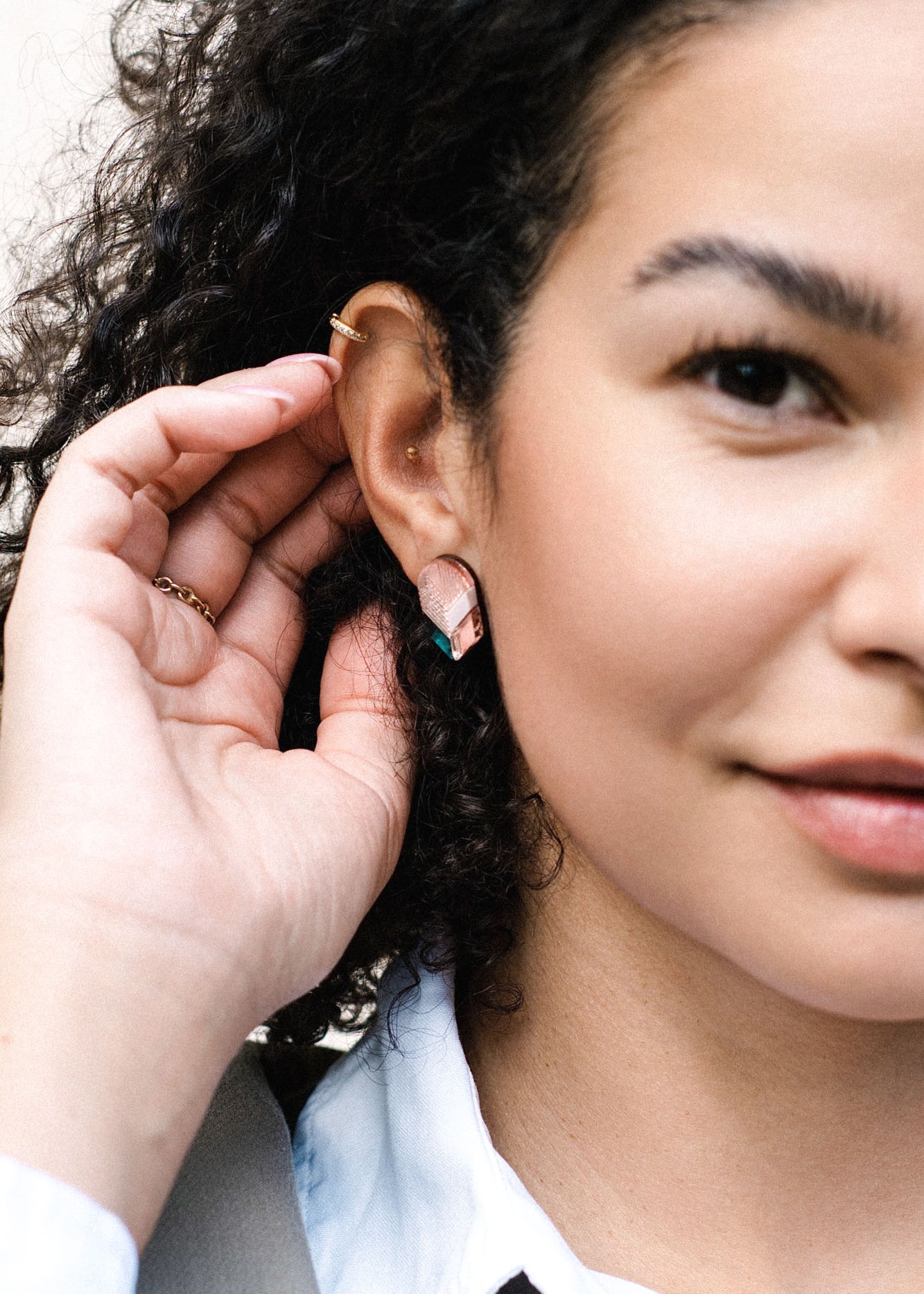 A close-up of a woman with curly hair touching her ear, showcasing minimalist jewelry with geometric earrings and a gold ear cuff. She is wearing a white collared shirt and light-colored jacket, smiling slightly.