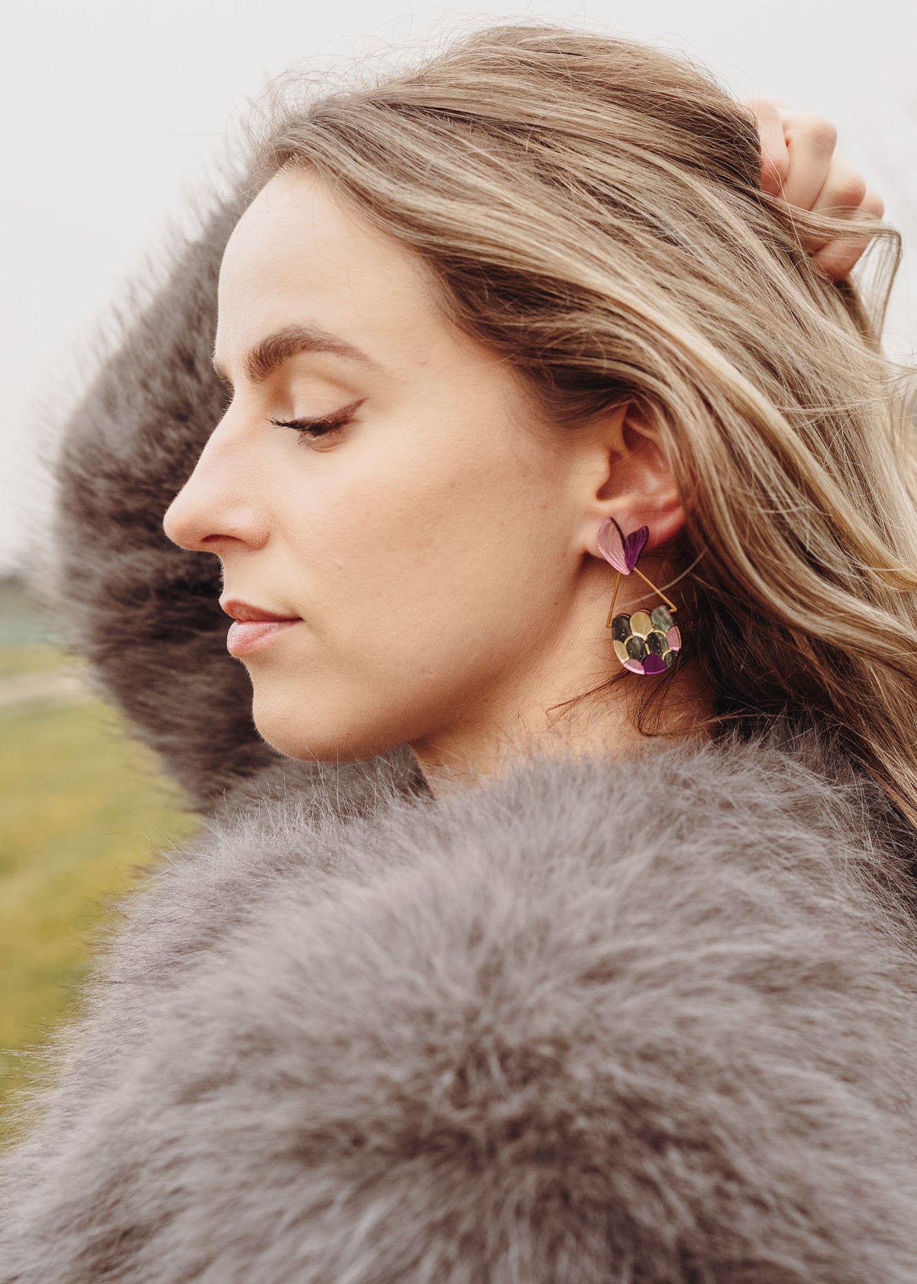 A woman with wavy blonde hair, wearing a fluffy gray coat and playful Mermaid Tail Earrings, stands outdoors with her eyes closed and one hand in her hair. The background is grassy with blurred trees—perfect for mermaid lovers.