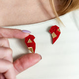 A person with long hair wears a cream top, holding a red and gold geometric One Love Halves Pin shaped like half a broken heart—the other handcrafted half is pinned to their shirt near the collarbone. Perfect as a romantic gift.