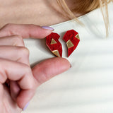 A person wearing a white top holds a red, geometric, handcrafted One Love Halves Pin against their chest. The heart-shaped pin is split into two pieces with gold accents, making it a meaningful romantic gift.