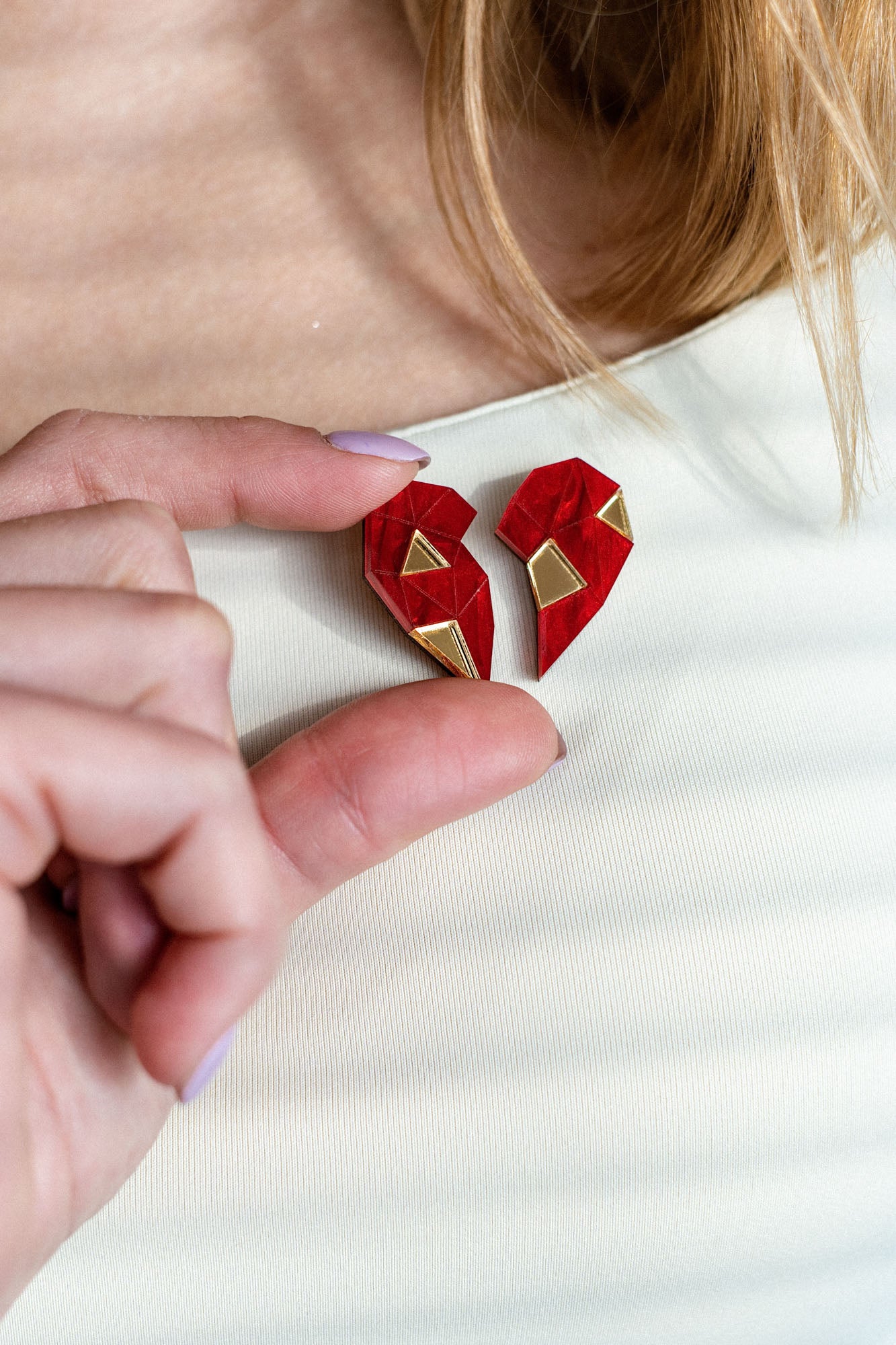 A person wearing a white top holds a red, geometric, handcrafted One Love Halves Pin against their chest. The heart-shaped pin is split into two pieces with gold accents, making it a meaningful romantic gift.