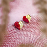 A pair of handcrafted red and gold geometric heart-shaped earrings rest on a textured pink fabric, with blurry plant stems in the foreground—perfect romantic jewelry for any occasion.