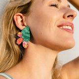 A woman with blonde hair smiles in sunlight, wearing a large, statement earring shaped like a green leaf with pink and orange accents, showcasing a bold design inspired by the opuncia cactus.