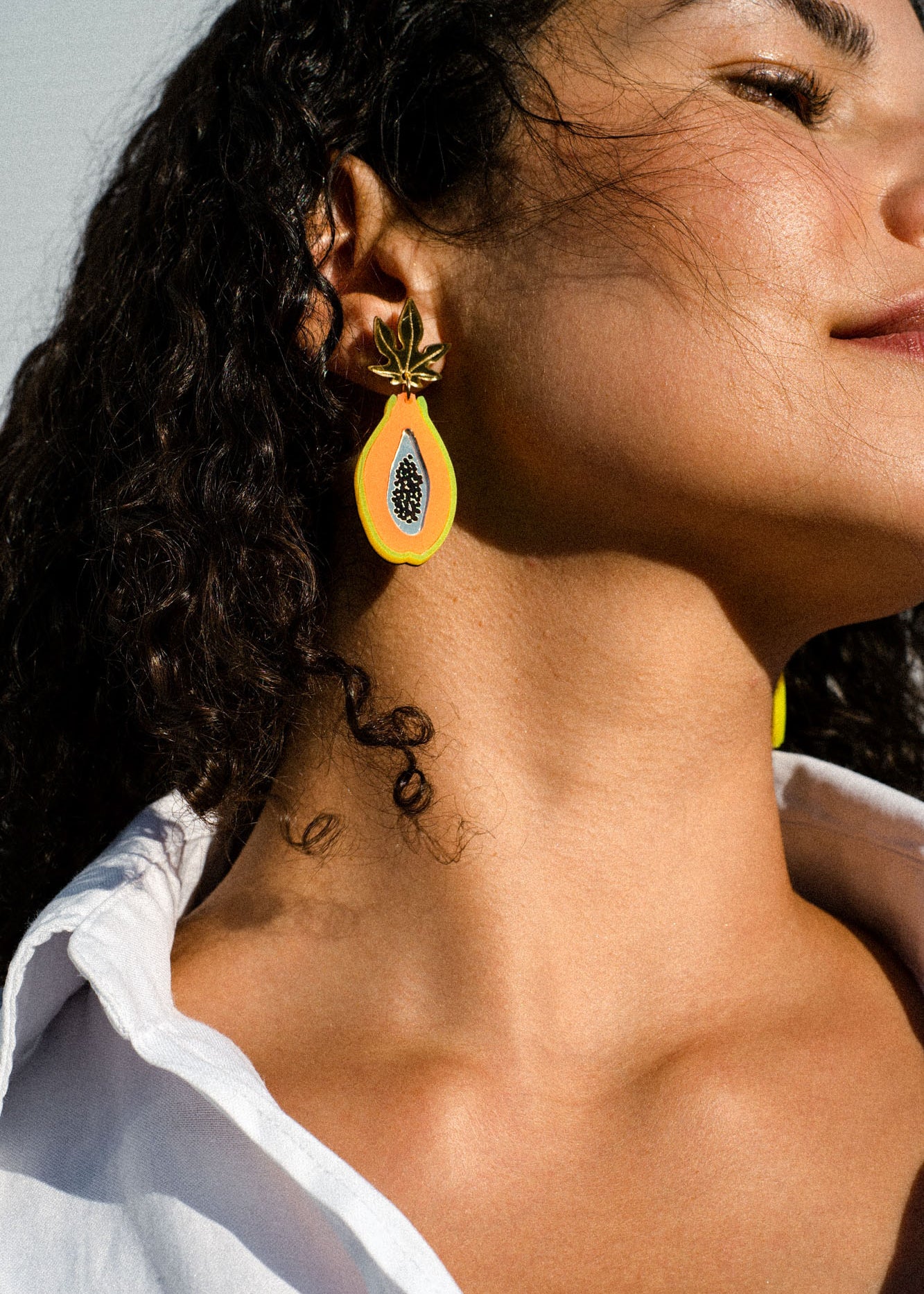 A woman with curly dark hair wears a white shirt and a colorful pair of Papaya Earrings, looking upward against a light background. These vibrant, handcrafted earrings add a playful touch to her modern style.
