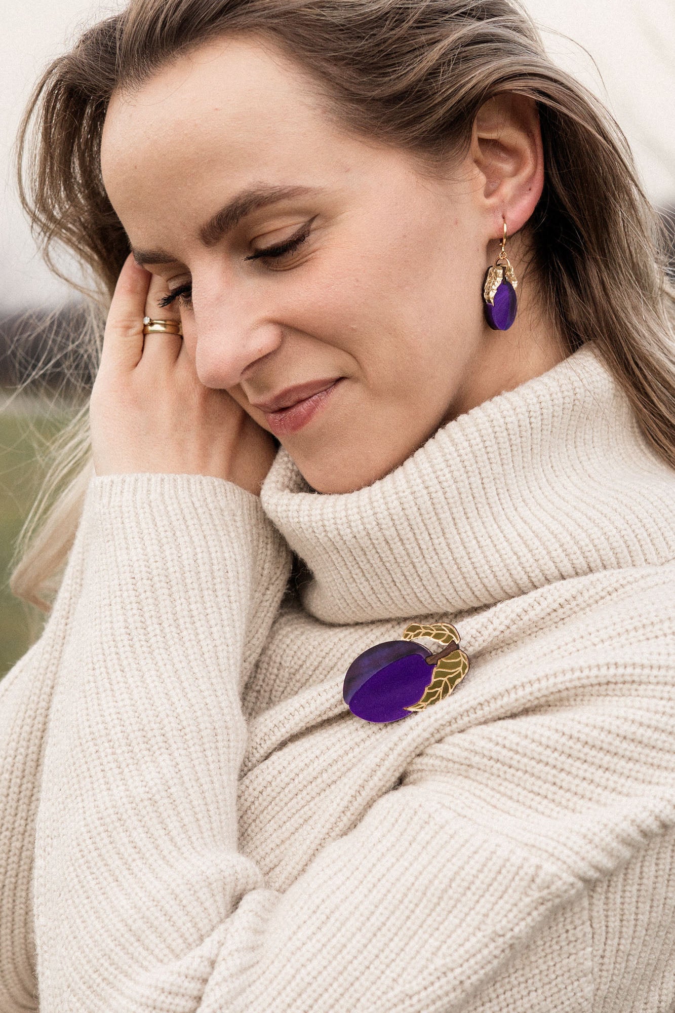 A woman wearing a cream-colored turtleneck sweater stands outdoors, smiling gently with eyes closed. She has wavy hair and wears matching purple jewelry—a handcrafted brooch and earrings. The grassy background is slightly blurred.