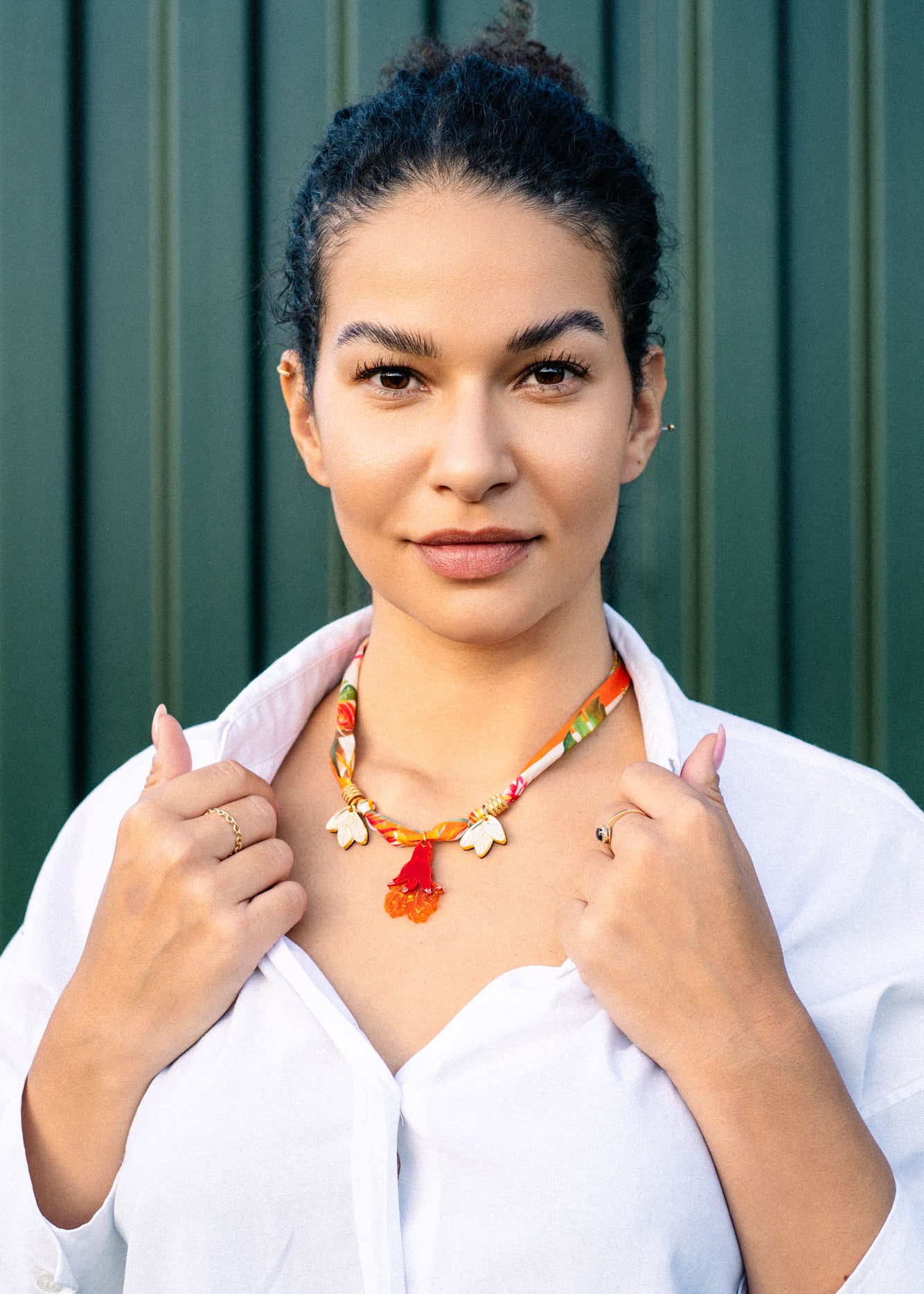 A woman with dark hair in a bun wears a white shirt and a handmade pomegranate flower necklace featuring floral charms on a silk-print cord, standing confidently before a green striped background while holding her collar.