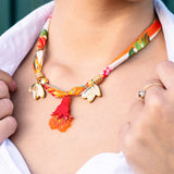 A woman wearing a handmade pomegranate flower necklace with gold accents, red tassels, and orange flower-shaped charms, paired with a white collared shirt. Only her lower face, neck, and hand are visible.