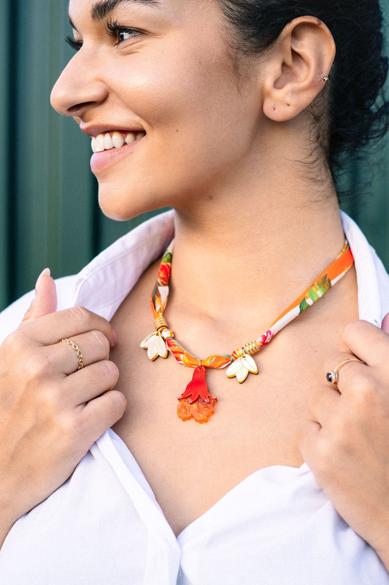 A woman smiling, wearing a white shirt and holding a handmade pomegranate flower necklace with gold and orange flower-shaped charms. She has small hoop earrings, rings on her fingers, and the necklace features a silk-print cord.