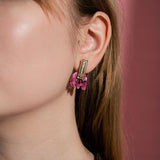 A close-up of a person’s ear wearing handcrafted gold and pink Romantic Tulip Earrings, shaped like a rectangle with dangling details, against a pink background. Only half of the person’s face is visible, highlighting these delicate, allergy-free earrings.