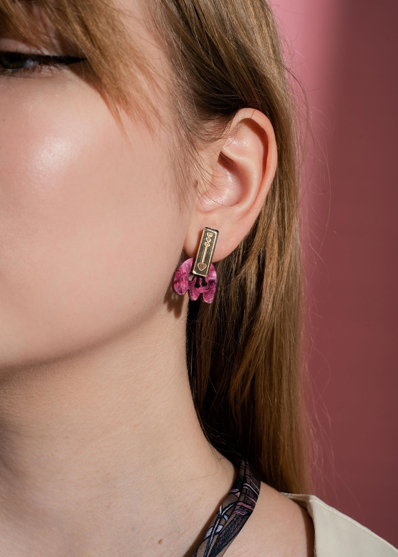 A close-up of a person’s ear wearing handcrafted gold and pink Romantic Tulip Earrings, shaped like a rectangle with dangling details, against a pink background. Only half of the person’s face is visible, highlighting these delicate, allergy-free earrings.