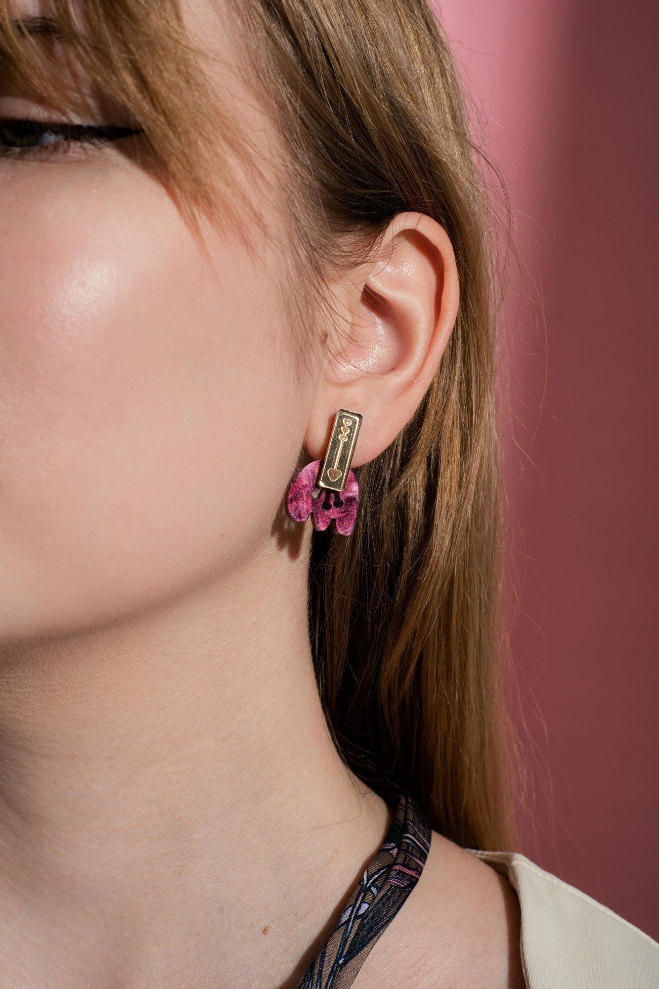A close-up of a person’s ear wearing handcrafted gold and pink Romantic Tulip Earrings, shaped like a rectangle with dangling details, against a pink background. Only half of the person’s face is visible, highlighting these delicate, allergy-free earrings.