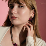 A young woman with long light brown hair poses against a pink background, wearing a cream top, winged eyeliner, and allergy-free handcrafted earrings—a dark necklace with a pink pendant and matching Romantic Tulip Earrings—gently touching her neck.