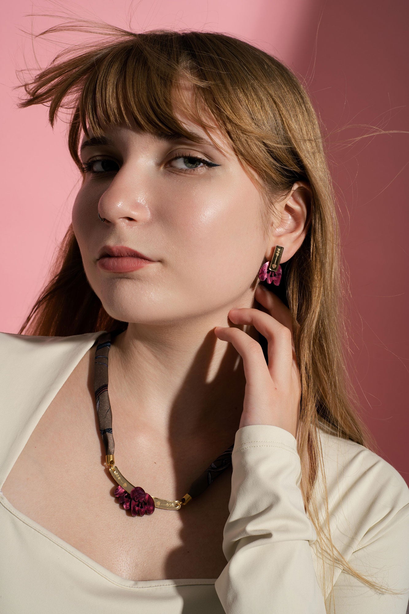 A young woman with long light brown hair poses against a pink background, wearing a cream top, winged eyeliner, and allergy-free handcrafted earrings—a dark necklace with a pink pendant and matching Romantic Tulip Earrings—gently touching her neck.