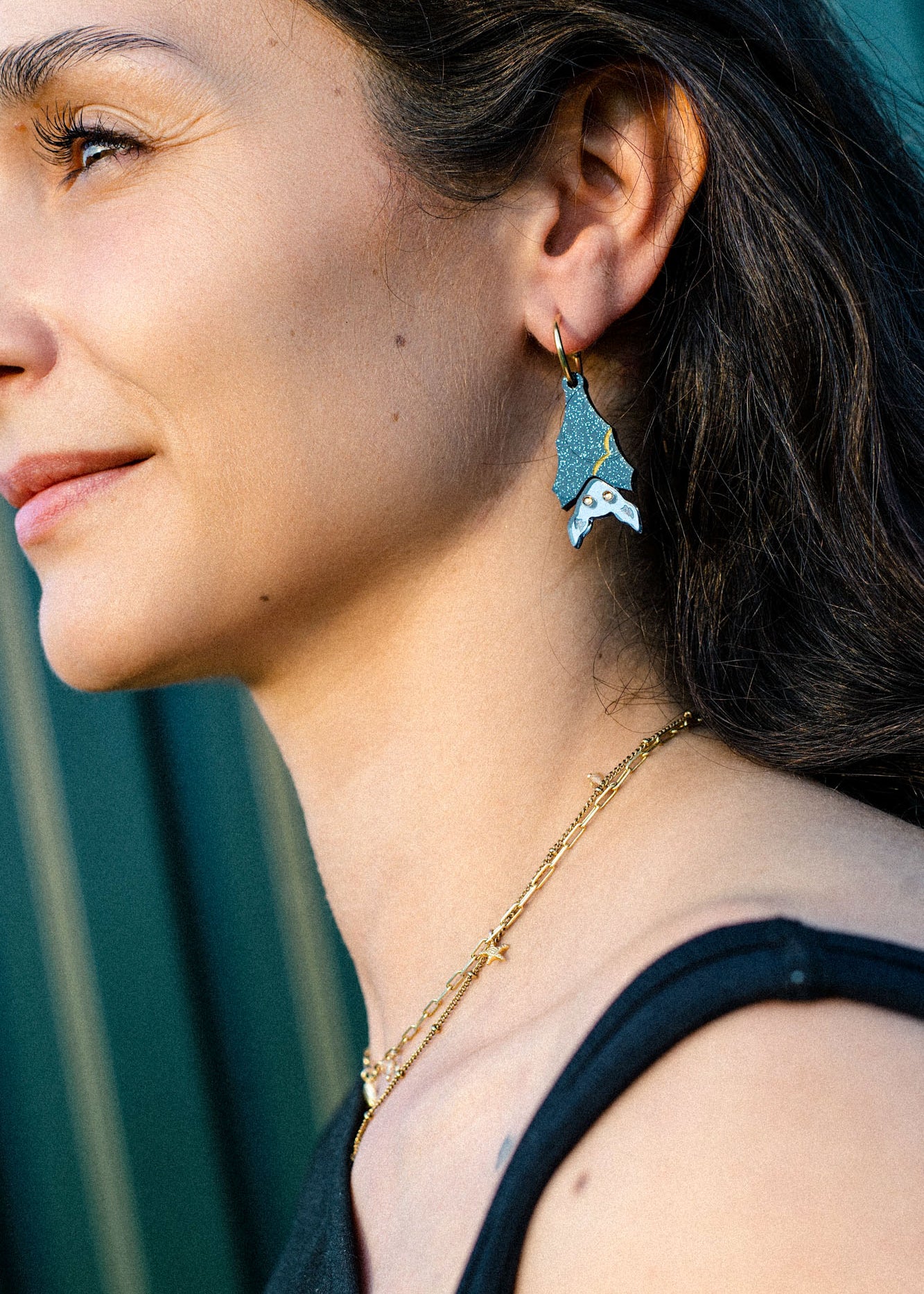 A woman with long brown hair wears a gold chain necklace and geometric, multi-colored earrings shaped like a fish. She is smiling slightly and looking to the left, sunlight highlighting her face in a subtle gothic glam style.