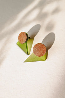 A pair of minimalist triangle earrings with round walnut wood tops and green acrylic glass bases are displayed on a light textured surface, casting soft shadows.