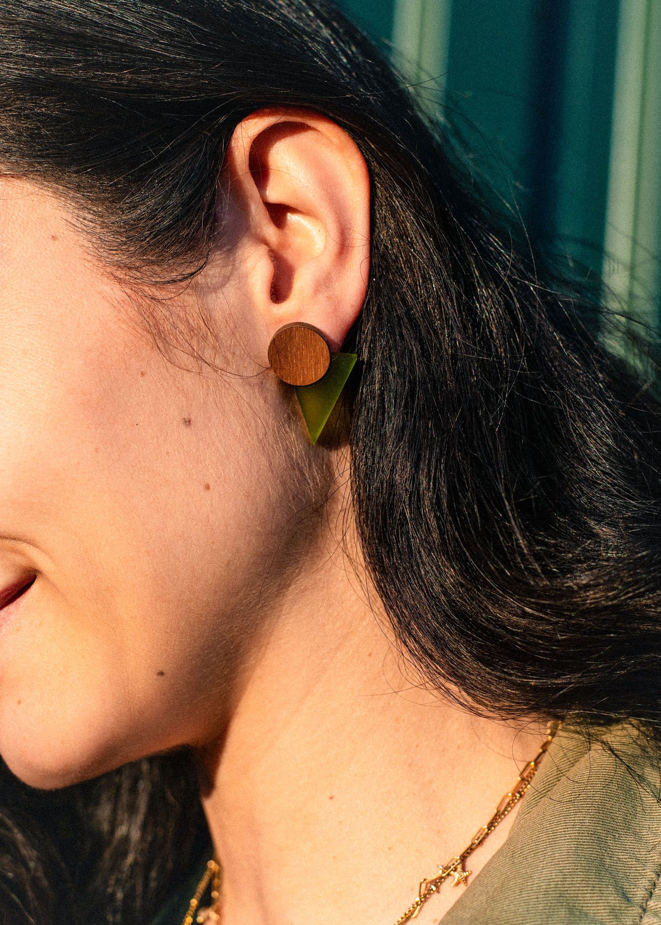 Close-up of a person with dark hair wearing striking triangle earrings crafted from acrylic glass, paired with a gold chain necklace. The background features blurred green vertical lines, adding to the modern aesthetic.