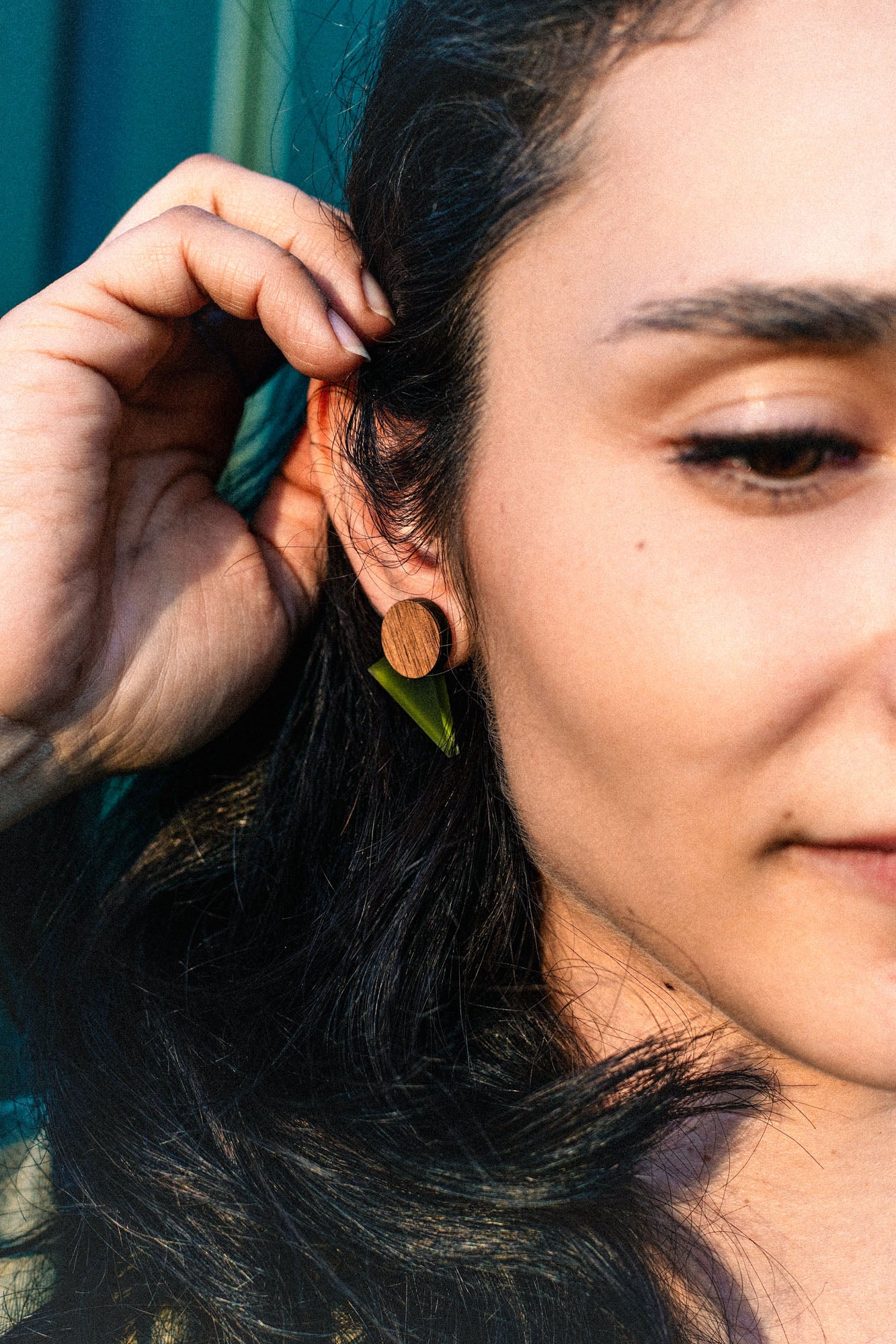 Close-up of a person with long dark hair touching their ear, wearing a round wooden earring and a small green leaf, sunlight highlighting their face and hand—a harmonious touch that's perfect for a capsule wardrobe.