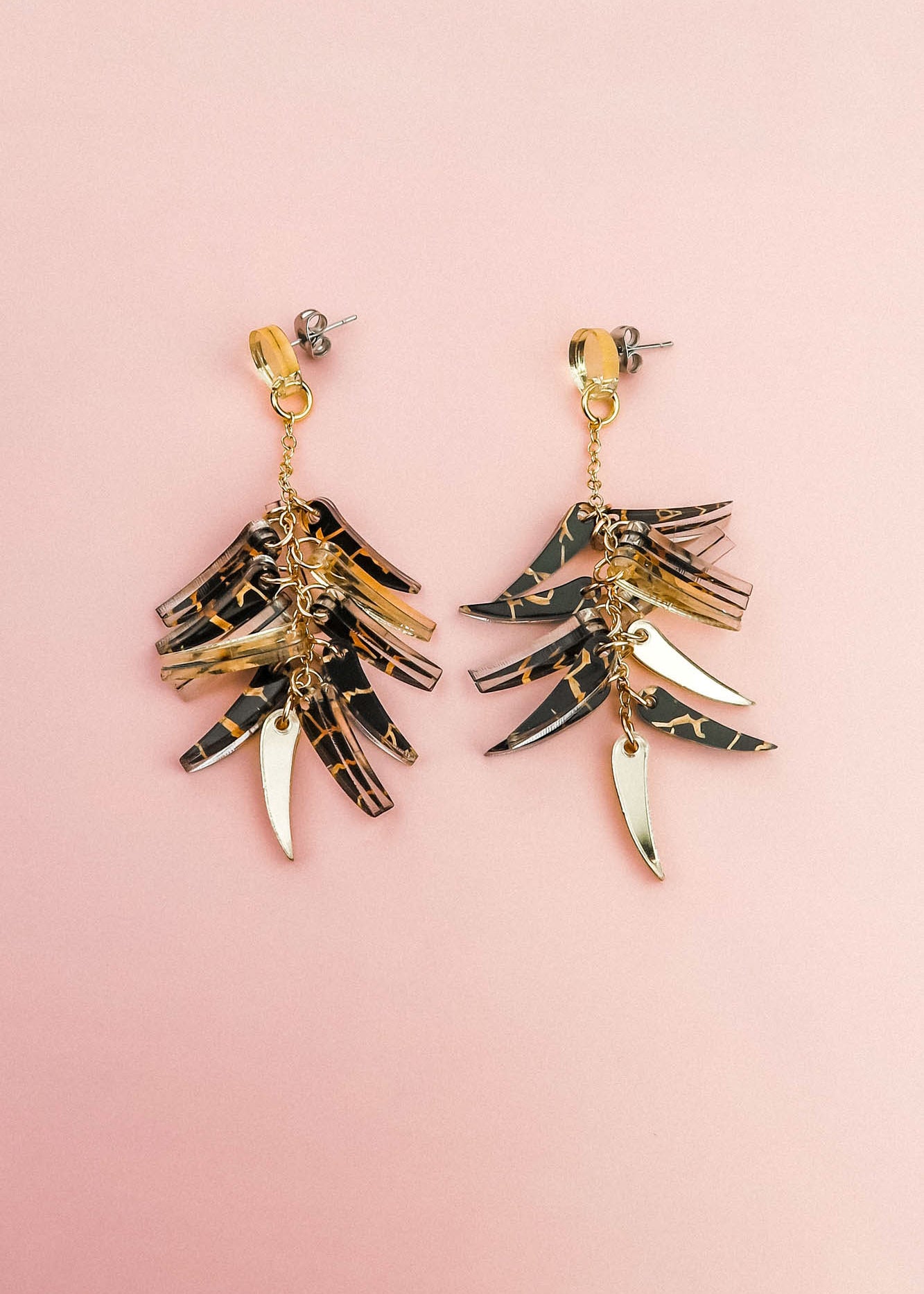 A pair of featherlight dangle earrings with layered gold and tortoiseshell-patterned pieces arranged in a fan-like design, displayed on a light pink background.