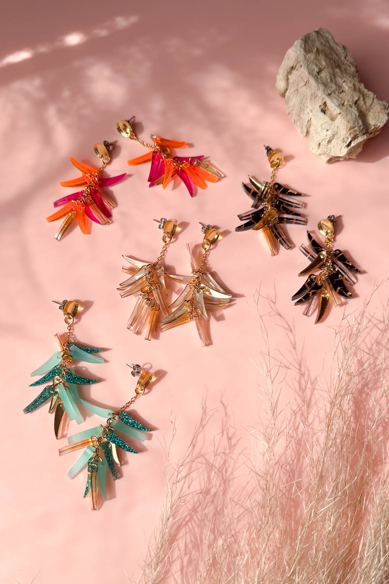 Colorful featherlight dangle earrings in four pairs—red-orange, brown-black, teal, and beige—feature acrylic tufts and are arranged on a pink surface with natural light, a beige rock, and wispy dried grass in the background.